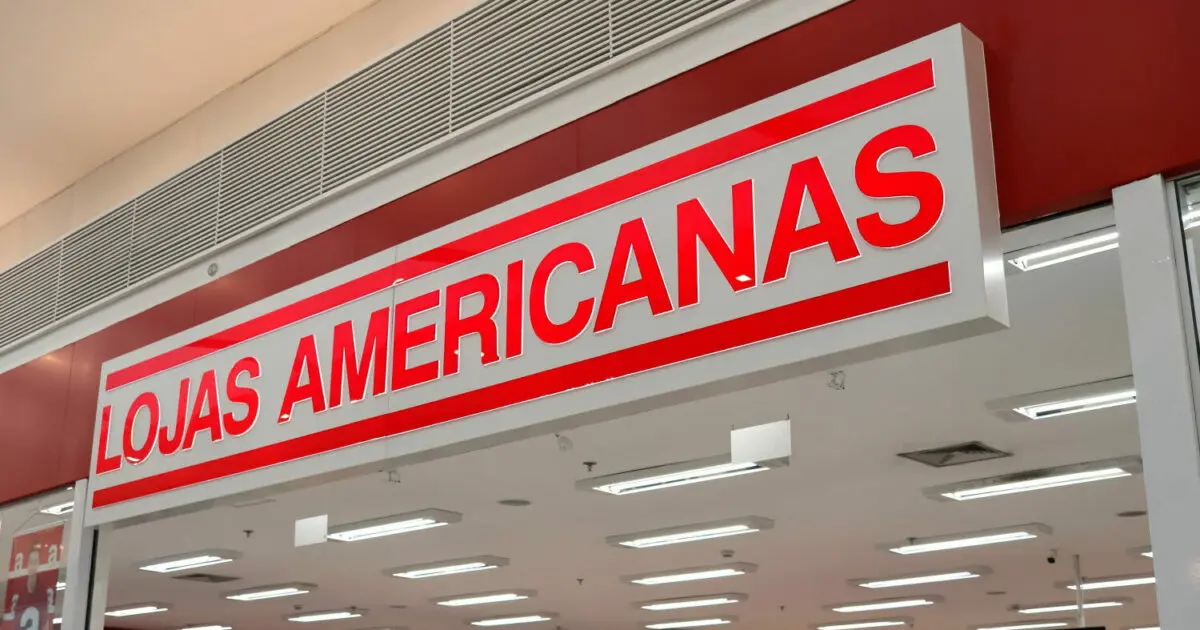 Fachada de uma loja da rede Lojas Americanas, com um letreiro branco e vermelho escrito em letras grandes e destacadas "LOJAS AMERICANAS". A loja está localizada dentro de um ambiente fechado, com luminárias visíveis no teto e detalhes arquitetônicos ao redor (lista, recuperação judicial)