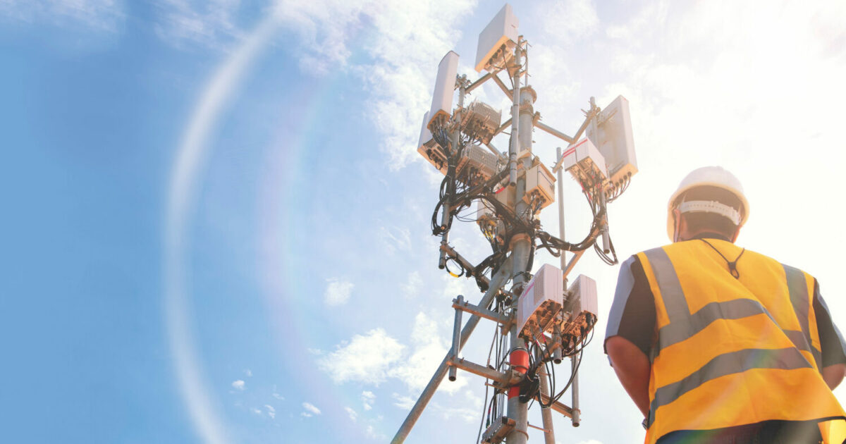 Imagem de um trabalhador usando colete de alta visibilidade e capacete, olhando para uma torre de telecomunicações equipada com antenas e equipamentos de transmissão. O céu está azul com algumas nuvens, iluminado pela luz do sol. A cena representa o avanço tecnológico e a infraestrutura necessária para a conectividade 5G (redes, Anatel, 5G)