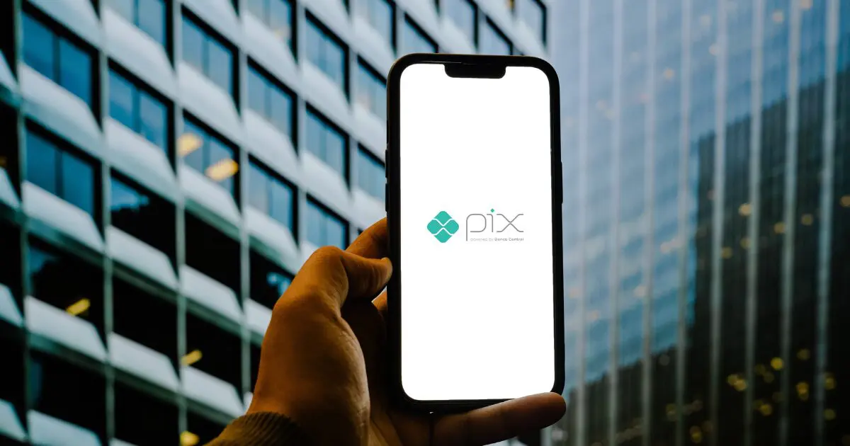 Pessoa segurando um smartphone com a tela exibindo o logotipo do Pix, sistema de pagamentos instantâneos do Banco Central do Brasil, tendo ao fundo prédios comerciais de vidro