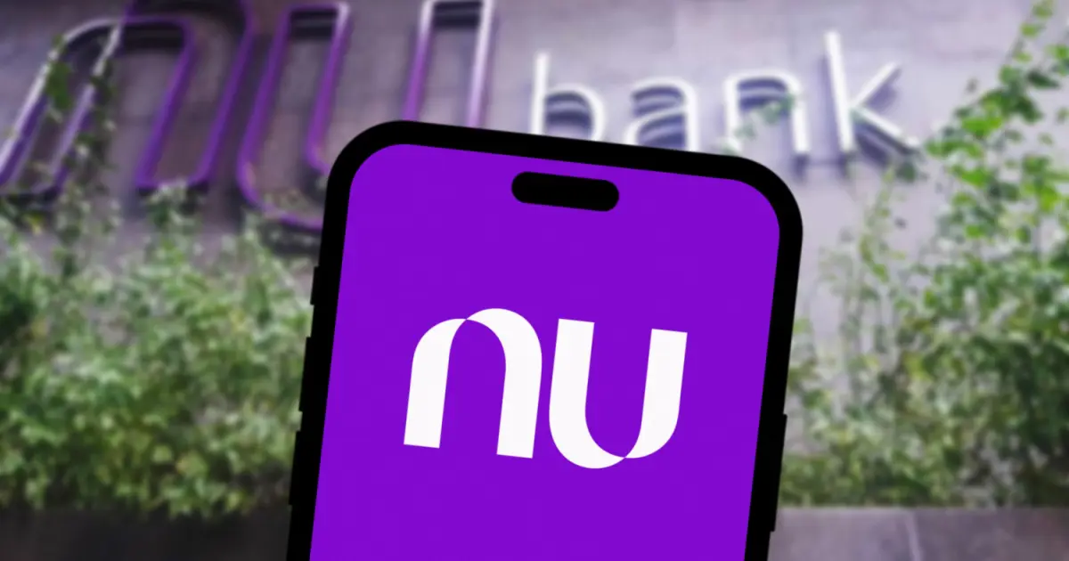 Imagem do logotipo do Nubank exibido em um dispositivo móvel com fundo roxo, enquanto ao fundo aparece a fachada de um prédio com o nome 'bank' parcialmente visível e cercado por vegetação