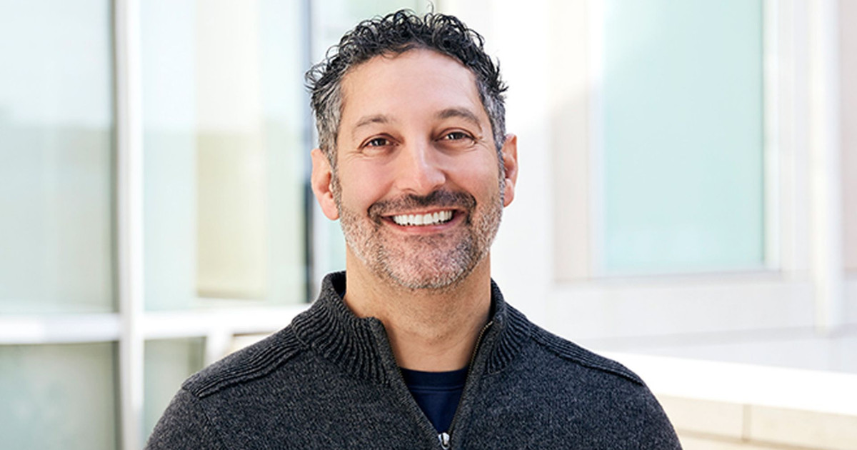 Retrato de Amit Yoran, CEO da Tenable, sorrindo em um ambiente iluminado por luz natural. Ele está vestindo um suéter cinza com zíper, e o fundo apresenta uma arquitetura moderna com janelas grandes que refletem luz suave. A imagem transmite uma sensação de profissionalismo e acessibilidade