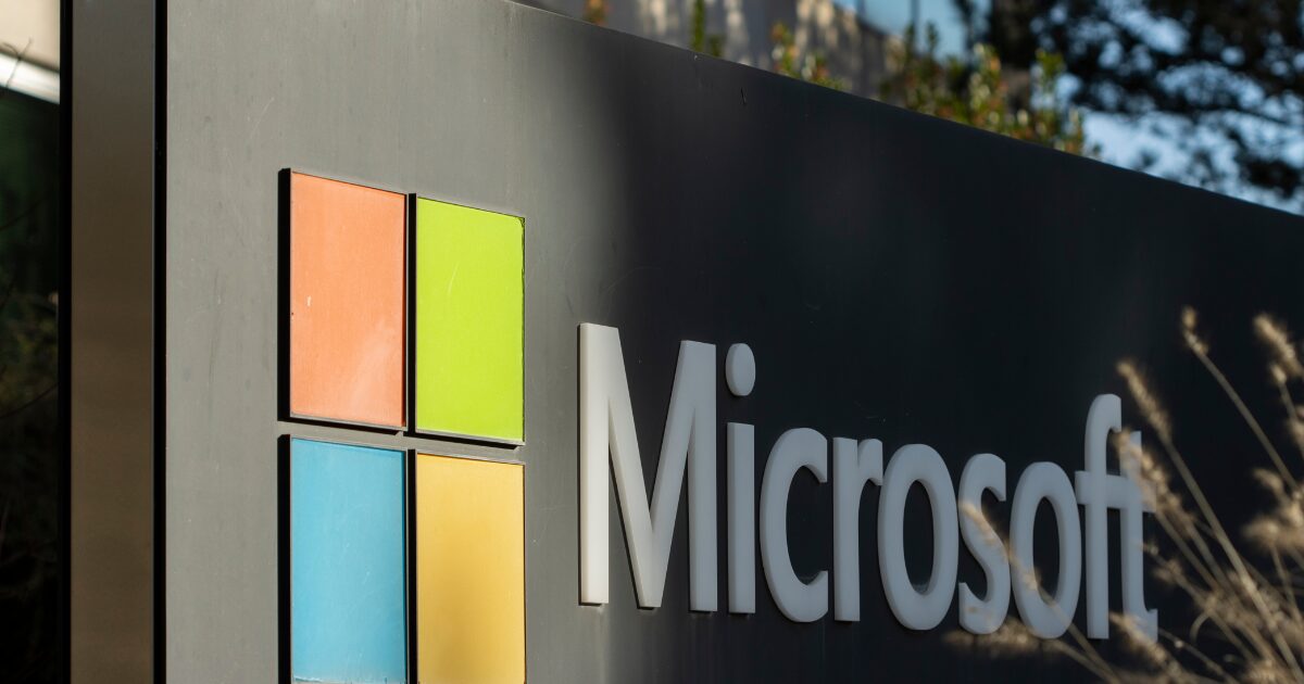 Placa externa da sede da Microsoft em um dia ensolarado, exibindo o logotipo da empresa com o símbolo de quatro quadrados coloridos (laranja, verde, azul e amarelo) à esquerda e o nome 'Microsoft' em letras brancas à direita. Ao fundo, folhagens e um prédio de vidro desfocado.