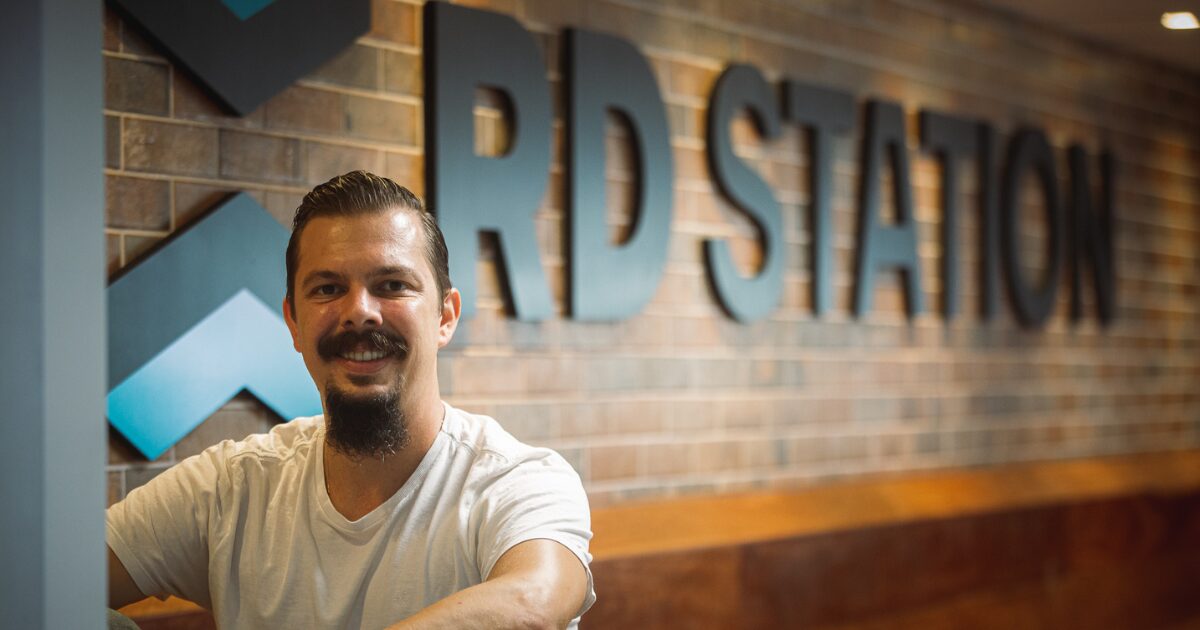 Imagem de um Bruno Ghisi sentado em frente a uma parede com o logo da RD Station. Ele usa uma camiseta branca e tem barba e bigode. O logo da empresa está em letras grandes e pretas sobre um fundo de tijolos, ao lado de um símbolo em tons de azul e preto. A iluminação é suave e destaca o rosto do homem, criando um ambiente descontraído e acolhedor