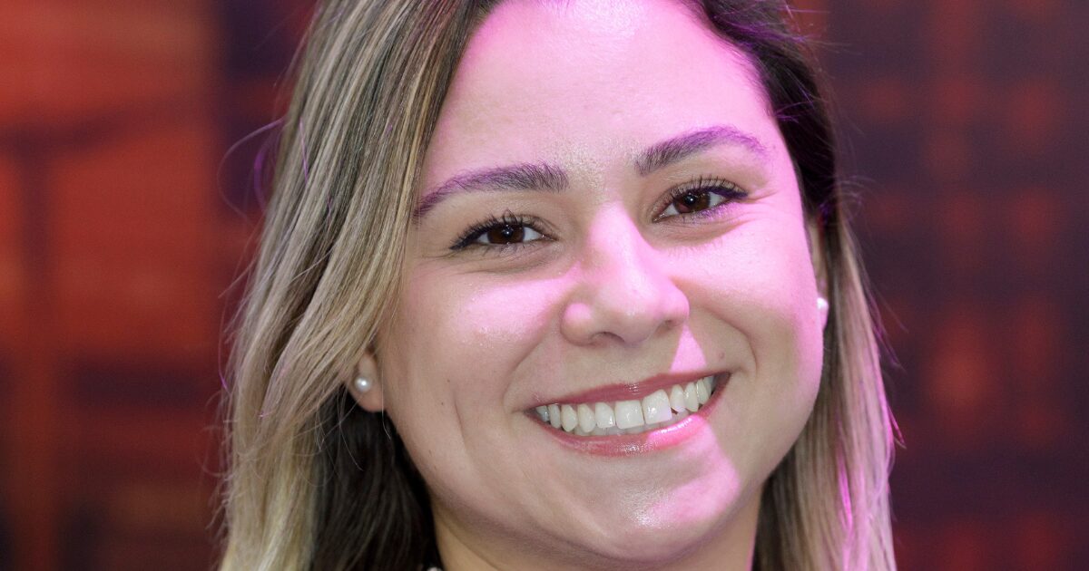 Uma mulher sorrindo com o rosto levemente iluminado por uma luz rosada. Ela tem cabelos lisos e claros, que caem sobre os ombros. Seus olhos são castanhos, e ela usa brincos de pérola. A imagem tem um fundo desfocado em tons quentes, destacando o rosto dela. Especialista da Oakmont