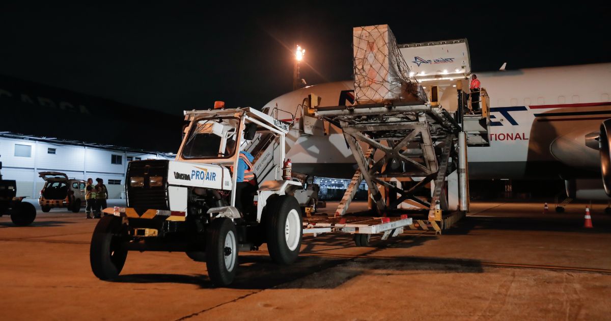 Carga do supercomputador sendo descarregada no Aeroporto do Galeão. A imagem mostra um trator de transporte de carga com a inscrição 'PRO AIR', movendo uma grande caixa de carga protegida por uma rede, com o avião de transporte visível ao fundo, em operação noturna sob iluminação artificial