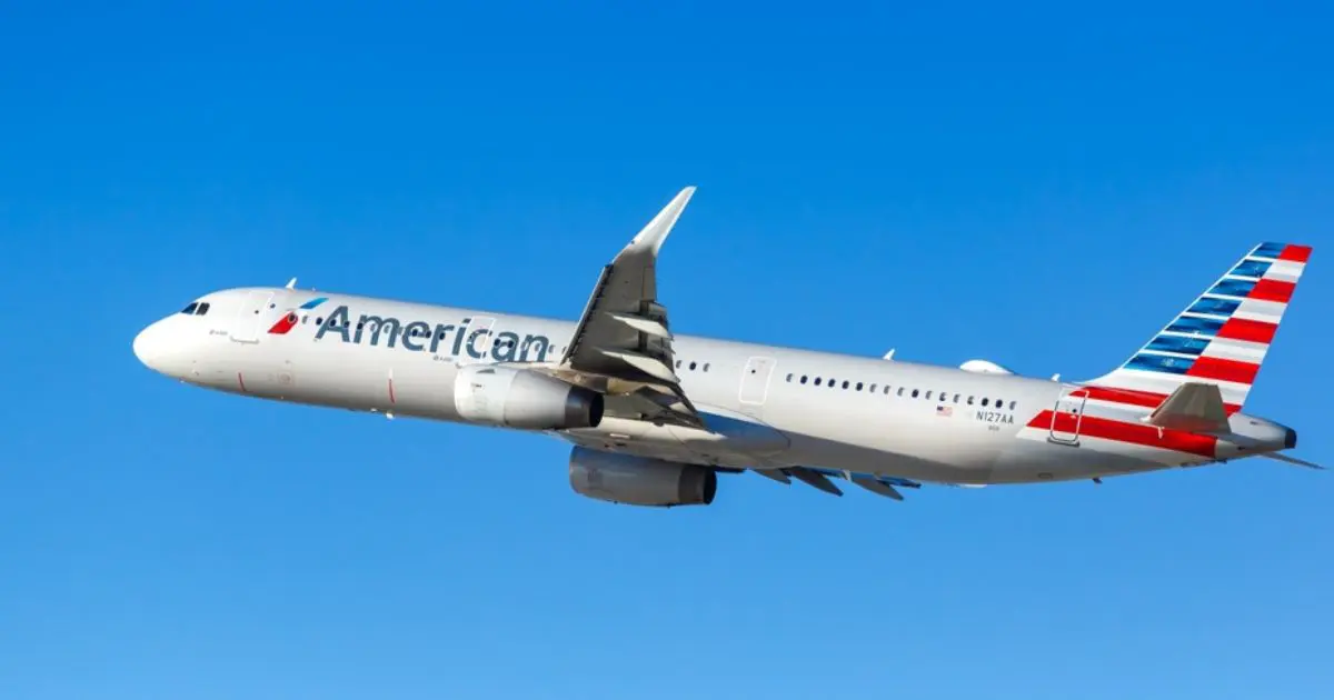 A imagem mostra uma aeronave da American Airlines em pleno voo, contra um céu claro e azul. O avião exibe as cores características da companhia, com sua fuselagem branca, o logotipo da American Airlines na lateral e o leme de cauda pintado com listras verticais vermelhas, brancas e azuis, simbolizando a bandeira dos Estados Unidos. A iluminação do dia destaca os detalhes do avião enquanto ele cruza os céus.