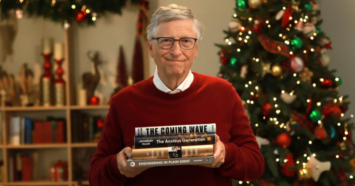 Bill Gates, vestindo um suéter vermelho, segura uma pilha de livros recomendados em frente a uma árvore de Natal decorada com luzes e enfeites. Ao fundo, uma estante com presentes e ornamentos natalinos completa o cenário festivo