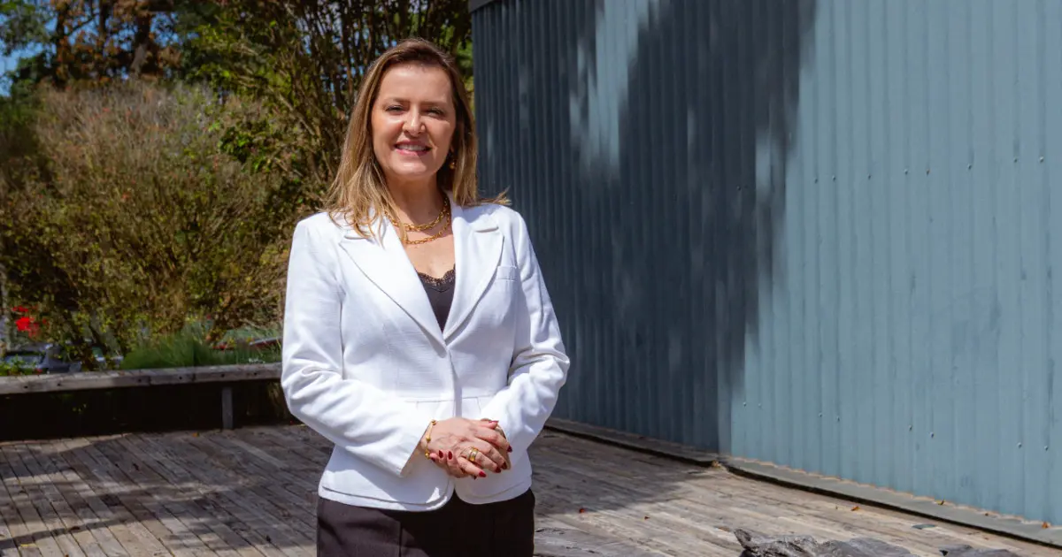 Cristiane Gomes, diretora de Tecnologia e Digital da CCR, em uma área externa com árvores ao fundo e uma parede azul. Ela está sorrindo, vestida com um blazer branco e camisa preta, transmitindo profissionalismo