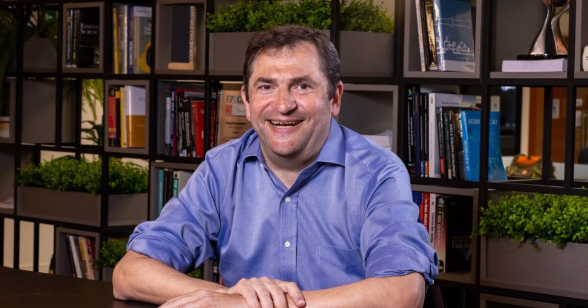 João Carlos Chachamovitz, CEO da Raxix, sorrindo em um escritório moderno com estantes cheias de livros e plantas ao fundo. Ele veste uma camisa azul e está sentado à mesa, transmitindo simpatia e profissionalismo