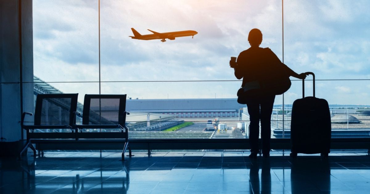 A imagem mostra uma cena típica de um aeroporto, com uma pessoa em silhueta olhando para um avião que decola ao fundo. A pessoa está próxima a uma janela de vidro, segurando uma xícara e com uma mala de rodinhas ao lado, sugerindo que está viajando ou esperando seu voo. O cenário inclui o terminal do aeroporto ao fundo, com pistas, hangares e um céu parcialmente nublado, iluminado pela luz do sol. A composição transmite uma sensação de movimento e transição, associada ao ato de viajar.