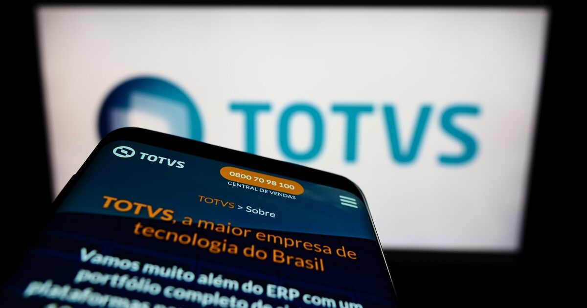 Um smartphone exibindo o site da Totvs, destacando o texto 'Totvs, a maior empresa de tecnologia do Brasil', com o logótipo da empresa ao fundo numa tela desfocada (reforma tributária)