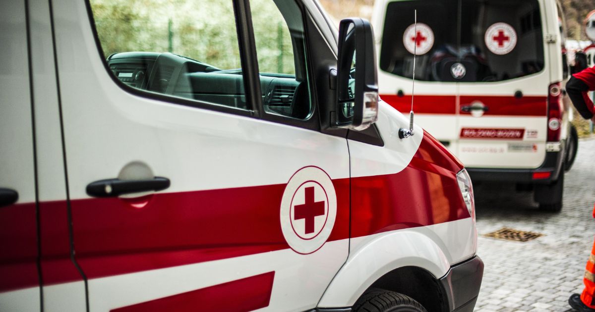 A imagem mostra duas ambulâncias de cor branca com uma faixa vermelha horizontal e o símbolo de uma cruz vermelha dentro de um círculo branco. No primeiro plano, vemos uma parte lateral e frontal da primeira ambulância, com a porta fechada e o vidro refletindo árvores ao fundo. No plano de fundo, há outra ambulância, parcialmente visível, estacionada em um pavimento de pedra. Ao lado direito da imagem, uma pessoa de uniforme laranja fluorescente aparece parcialmente, sugerindo uma equipe de resgate ou emergência.