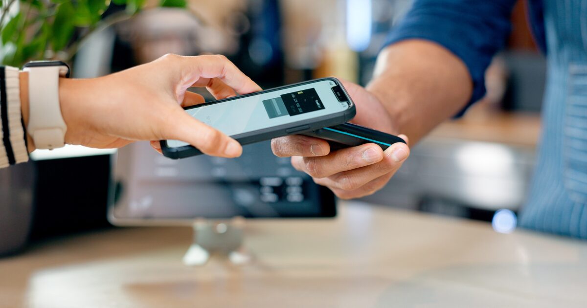 Uma pessoa realiza um pagamento digital por aproximação utilizando um smartphone. A cena ocorre em um ambiente comercial, com um dispositivo POS (point of sale) sendo usado para processar a transação. Ao fundo, desfocado, há uma tela de caixa registradora e detalhes do local.