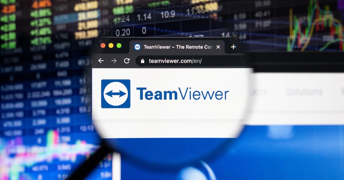 Imagem da interface do navegador de internet mostrando a página inicial do site da TeamViewer (teamviewer.com/en/), destacada por uma lupa sobre o logotipo da empresa, composto por um ícone azul com setas brancas e o texto "TeamViewer". Ao fundo, gráficos financeiros e números desfocados sugerem um contexto de negócios e tecnologia.