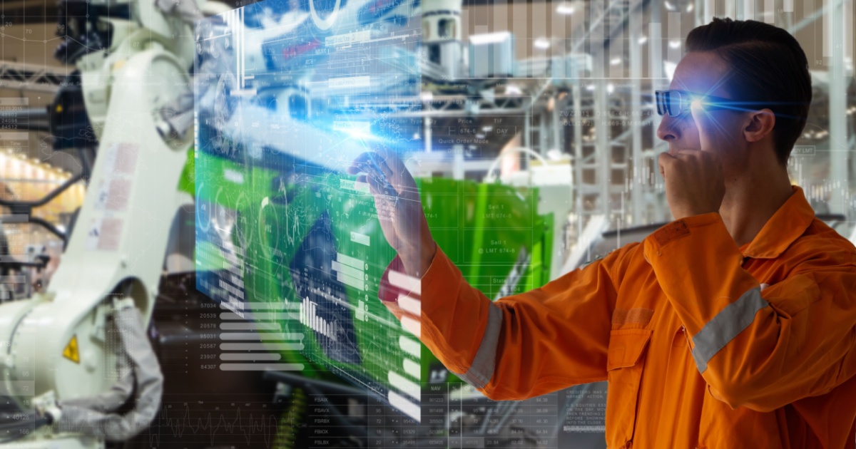 A imagem mostra um trabalhador de uniforme laranja interagindo com uma interface digital em um ambiente industrial. Ele usa óculos inteligentes e está manipulando gráficos e dados virtuais que flutuam diante dele, representando a tecnologia de gêmeos digitais. No fundo, pode-se ver um braço robótico industrial e uma linha de produção. A cena sugere o uso de tecnologias avançadas para monitoramento e controle de processos de manufatura (digital twin, gemeos digitais, indústria, fábrica, manufatura)