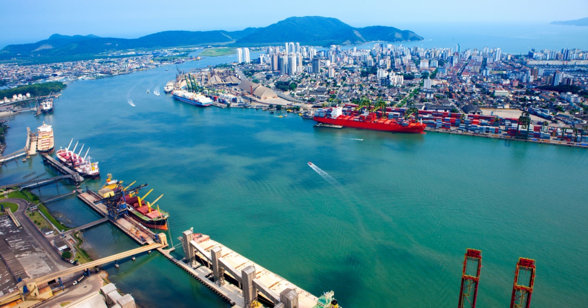 Vista aérea do Porto de Santos, no Brasil, mostrando navios atracados, guindastes e contêineres ao longo das margens do canal. Ao fundo, é possível ver a cidade de Santos com prédios e áreas urbanas, além de montanhas e o mar no horizonte (santos, porto de santos, santos brasil)