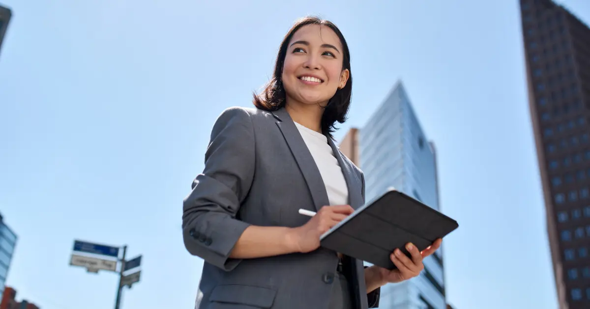 Mulher sorridente em ambiente corporativo ao ar livre, segurando um tablet e vestindo um terno cinza, com prédios modernos ao fundo sob um céu claro, vendas, capacitação