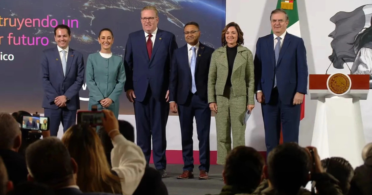 Evento de lançamento da infraestrutura de nuvem da AWS no México, com líderes e autoridades reunidos no palco. Entre os participantes estão Claudia Sheinbaum e outros representantes, todos em trajes formais. O fundo apresenta um painel com informações do evento e uma imagem aérea estilizada. À direita, há um pódio com o brasão do México e uma bandeira. O público na frente captura o momento com celulares, destacando a relevância do evento (AWS, Amazon Web Services)