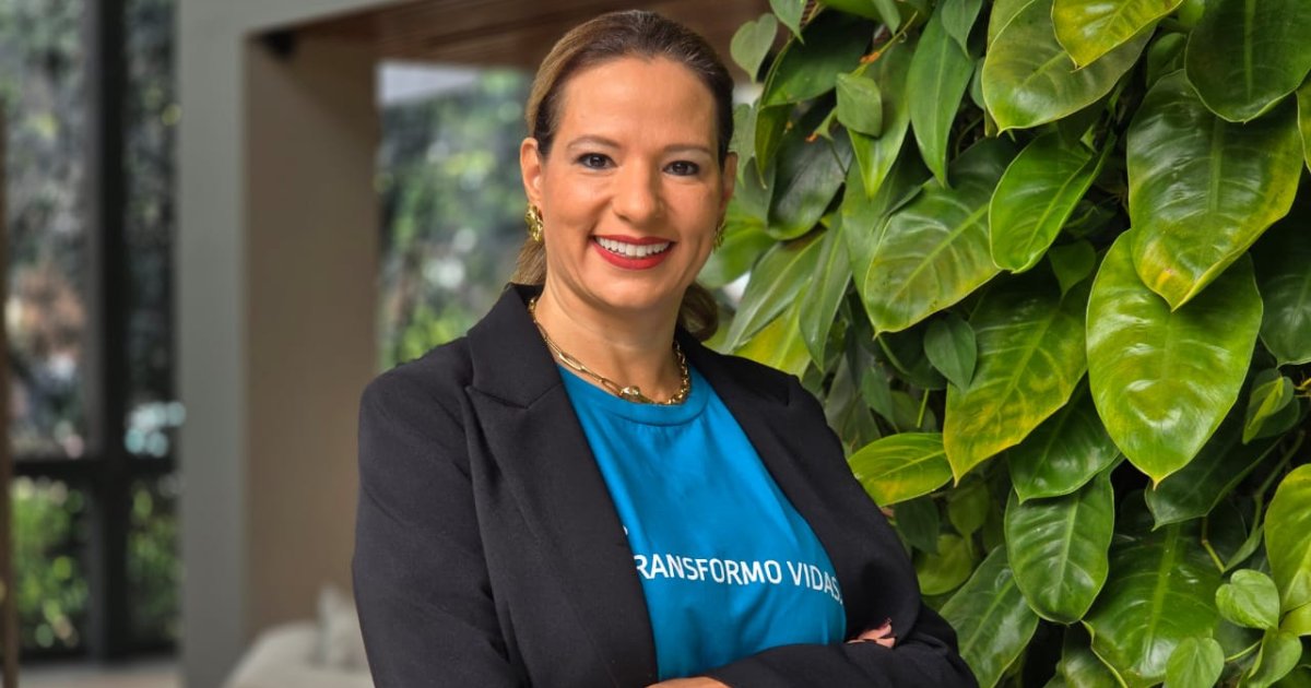 Ana Letícia Lucca, CRO da Escola da Nuvem, em destaque. Ela veste uma camiseta azul com a frase "Transformo Vidas" e um blazer preto. Ana está sorrindo e posando em frente a um painel de plantas verdes, com um fundo desfocado que revela um ambiente moderno e iluminado por luz natural. (Ana Letícia Lucca, Escola da Nuvem, foto Natália Rossi, formado)