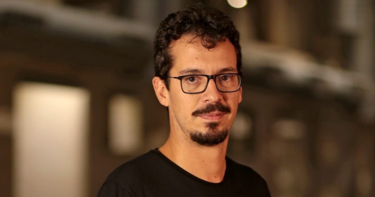 Gustavo Avelar, novo vice-presidente de Business Performance da Totvs, em destaque. Ele está usando uma camiseta preta e óculos de armação escura, com uma expressão confiante e o fundo desfocado, que sugere um ambiente interno com iluminação suave.