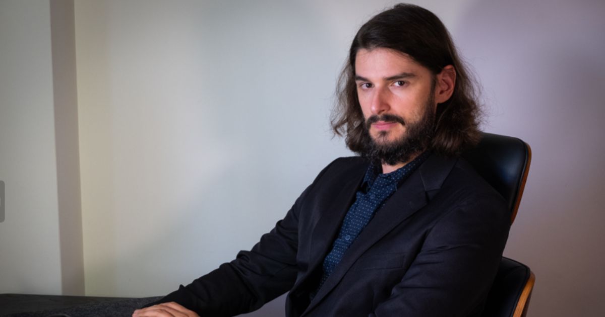 Na imagem, Lucas Rodrigues está sentado em uma cadeira de escritório, usando um terno escuro e uma camisa social azul escuro com padrão. Ele possui cabelos longos e barba cheia, aparentando estar em um ambiente de trabalho. Suas mãos estão repousando em um teclado de laptop, enquanto ele olha diretamente para a câmera, com uma expressão séria. O fundo da imagem é neutro e a iluminação destaca o sujeito, criando um tom profissional.