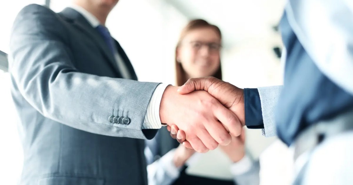 Dois executivos apertando as mãos em sinal de acordo, representando fusão e aquisição, com uma mulher ao fundo sorrindo (M&A, fusao, aquisicao, negocio, acordo, desafios)