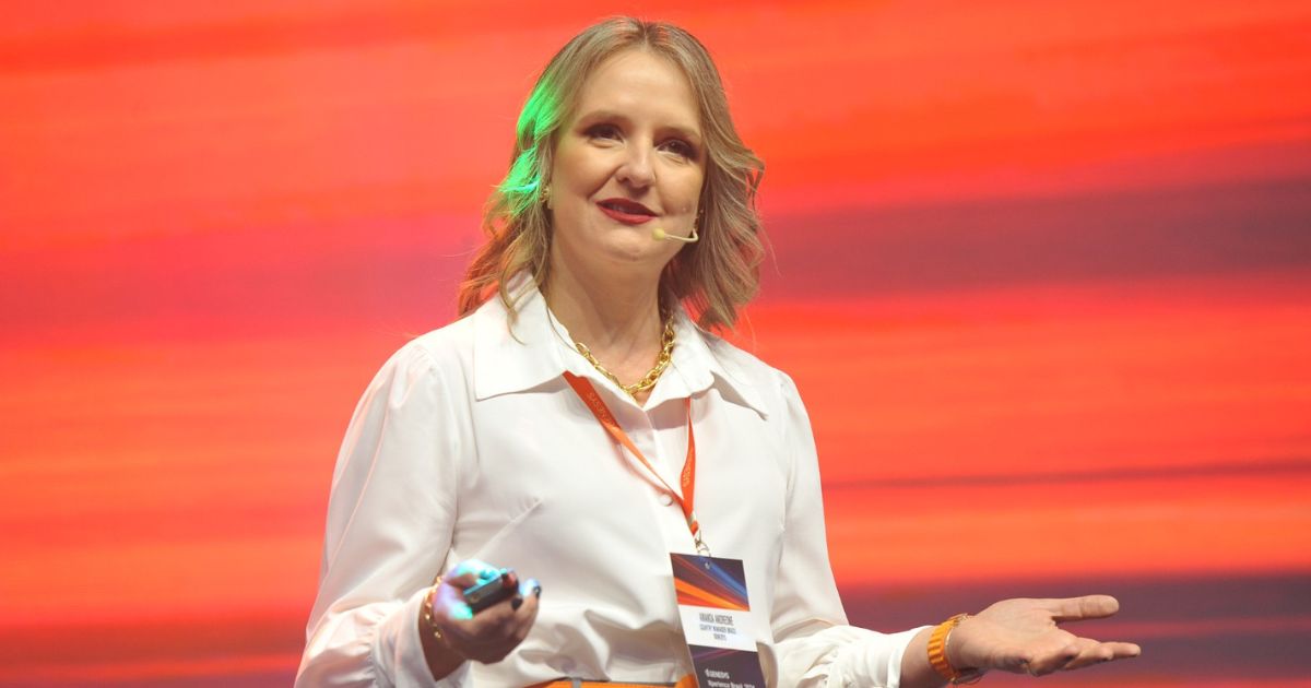 Amanda Andreone, Country Manager da Genesys Brasil, apresentando em um evento com fundo vibrante em tons de laranja e vermelho, usando camisa branca, calça cinza e acessórios em laranja.