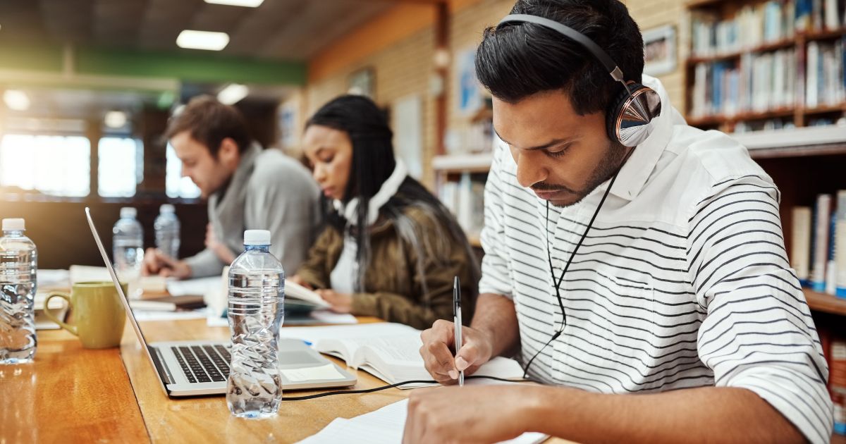 Três pessoas sentadas em uma mesa de madeira em uma biblioteca, estudando e escrevendo. Em primeiro plano, um homem com camisa listrada branca e preta, usando fones de ouvido, está concentrado escrevendo em um caderno. Há um laptop, uma garrafa de água e uma caneca na mesa à sua frente. Ao fundo, um homem e uma mulher também estão focados em leitura e escrita. A biblioteca tem estantes de livros ao fundo, e a luz natural entra pela janela, criando um ambiente iluminado e tranquilo