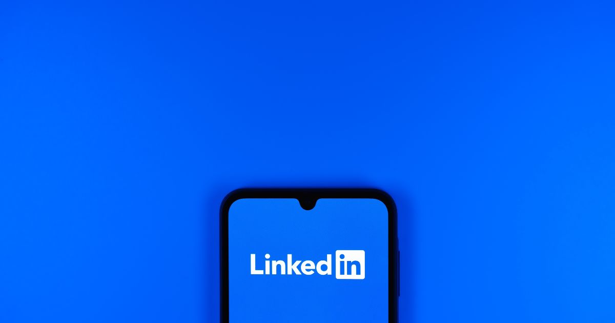 A imagem mostra um smartphone com a aplicação do LinkedIn aberta, destacada contra um fundo azul vibrante. O logo do LinkedIn está em branco e centralizado na tela do telemóvel, transmitindo uma sensação de tecnologia e conectividade.