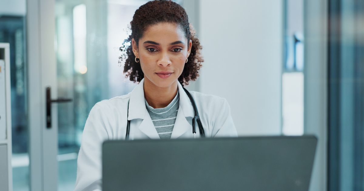 Uma médica jovem com cabelo encaracolado e preso para trás, usando um jaleco branco e estetoscópio ao redor do pescoço. Ela está sentada e parece concentrada enquanto olha para um computador. O ambiente parece ser um consultório ou hospital, com fundo claro e moderno.