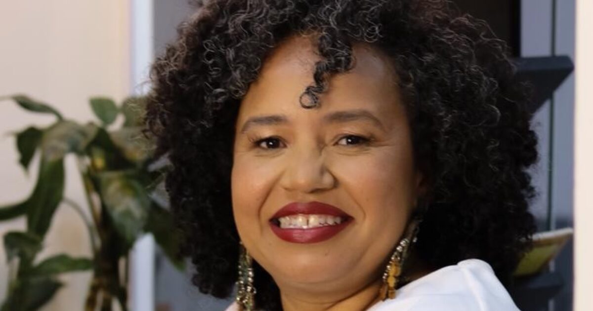 Fotografia de uma mulher sorrindo, com cabelos cacheados e pretos, usando batom vermelho. Ela veste uma blusa clara e brincos longos, e há plantas e uma estante ao fundo. Rosi Teixeira, nova CIO da Zup