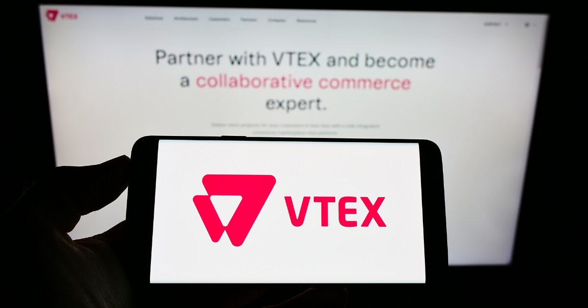 Logotipo da VTEX exibido na tela de um smartphone, com o site oficial da empresa ao fundo. No site, é possível ler a frase “Faça parceria com a VTEX e torne-se um especialista em comércio colaborativo”. A imagem destaca a identidade visual da marca e sua proposta de cola