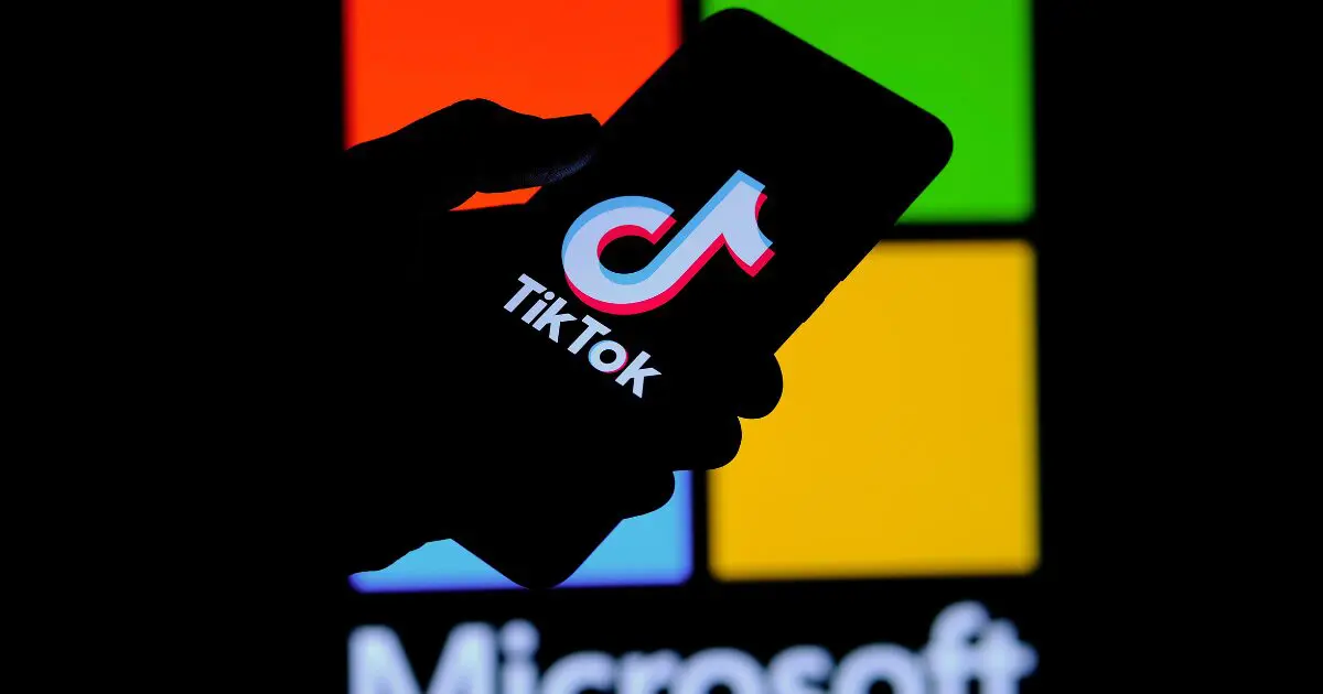 A imagem mostra a silhueta de uma mão segurando um smartphone, onde a tela exibe o logotipo do TikTok. Ao fundo, desfocado, aparece o logotipo da Microsoft com seus tradicionais blocos coloridos (vermelho, verde, azul e amarelo). A composição sugere uma relação entre as duas empresas, possivelmente indicando uma negociação, parceria ou aquisição.