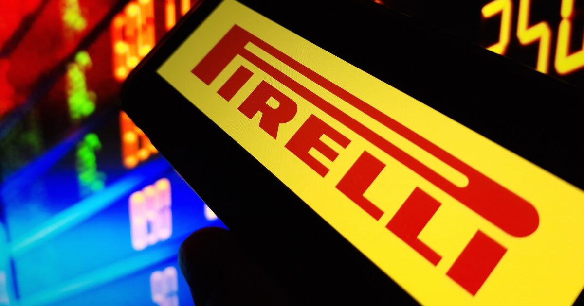 Tela de um smartphone exibe o logotipo da Pirelli em vermelho sobre um fundo amarelo, com um painel digital ao fundo mostrando números iluminados em tons de laranja e azul. A imagem transmite um ambiente tecnológico e de mercado financeiro.