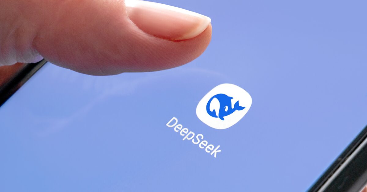 A imagem mostra um close-up de um dedo prestes a tocar o ícone do aplicativo "DeepSeek" na tela de um smartphone. O ícone tem um fundo branco com o desenho de uma baleia azul estilizada, e o nome "DeepSeek" aparece logo abaixo. A tela do dispositivo tem um fundo azul claro, criando um contraste suave (deepseek)