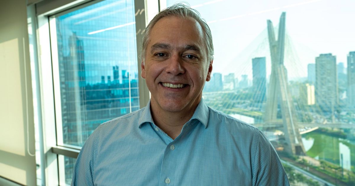 Homem de meia-idade sorrindo, vestindo uma camisa azul clara de botões, posicionado em frente a uma janela. Ao fundo, pode-se ver uma paisagem urbana com prédios altos e a Ponte Estaiada, um famoso marco arquitetônico.