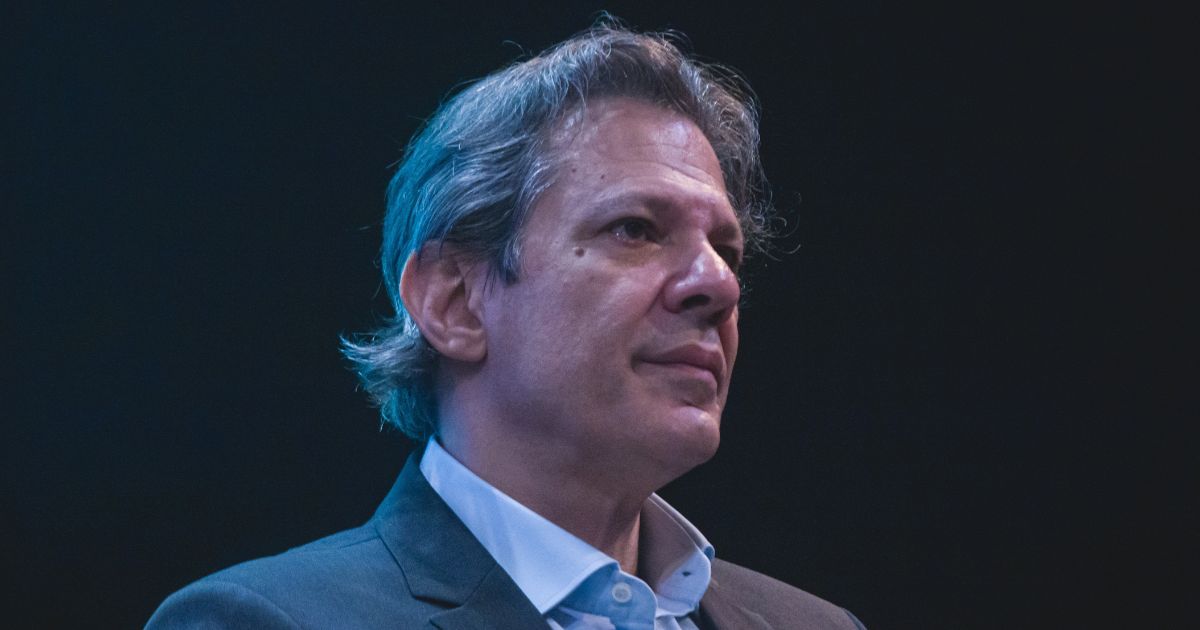 Esta imagem mostra um homem com cabelos grisalhos e expressão séria. Ele está vestido com um fato cinzento e camisa branca, olhando para o lado direito. O fundo é escuro, destacando seu rosto iluminado. (haddad, big tech, brasil)