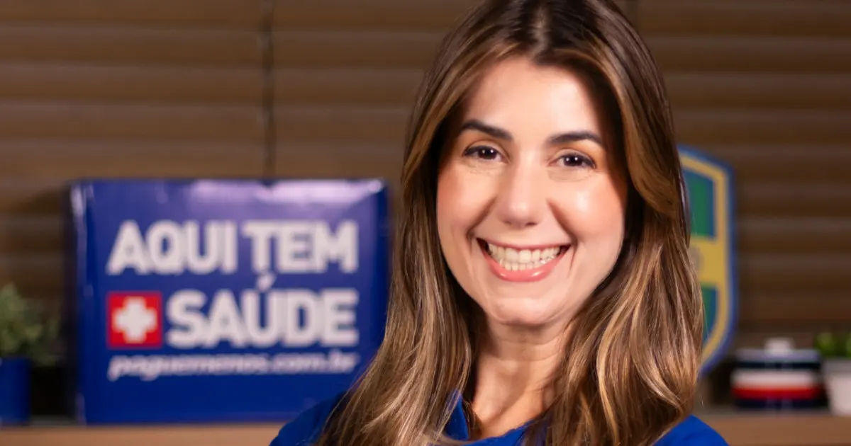 Priscila Braga, nova diretora de Digital da Pague Menos, sorrindo em um ambiente corporativo. Ao fundo, um cartaz azul com os dizeres 'Aqui tem saúde' reforça a identidade da empresa