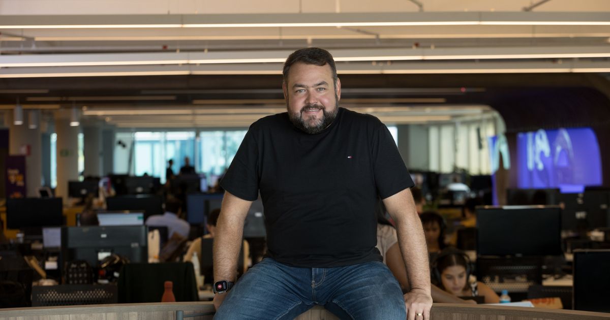 Na foto, vemos Taiolor Morais, diretor de Operações e Tecnologia da Youse, sentado de maneira descontraída em uma bancada dentro de um ambiente de escritório moderno e amplo. Ele veste uma camiseta preta de manga curta e calça jeans, mantendo os braços apoiados ao seu lado. Taiolor está sorrindo levemente e olha diretamente para a câmera, transmitindo uma expressão de confiança e simpatia. Ao fundo, observam-se diversas estações de trabalho e pessoas focadas em suas atividades, evidenciando o clima dinâmico de um grande escritório de tecnologia.