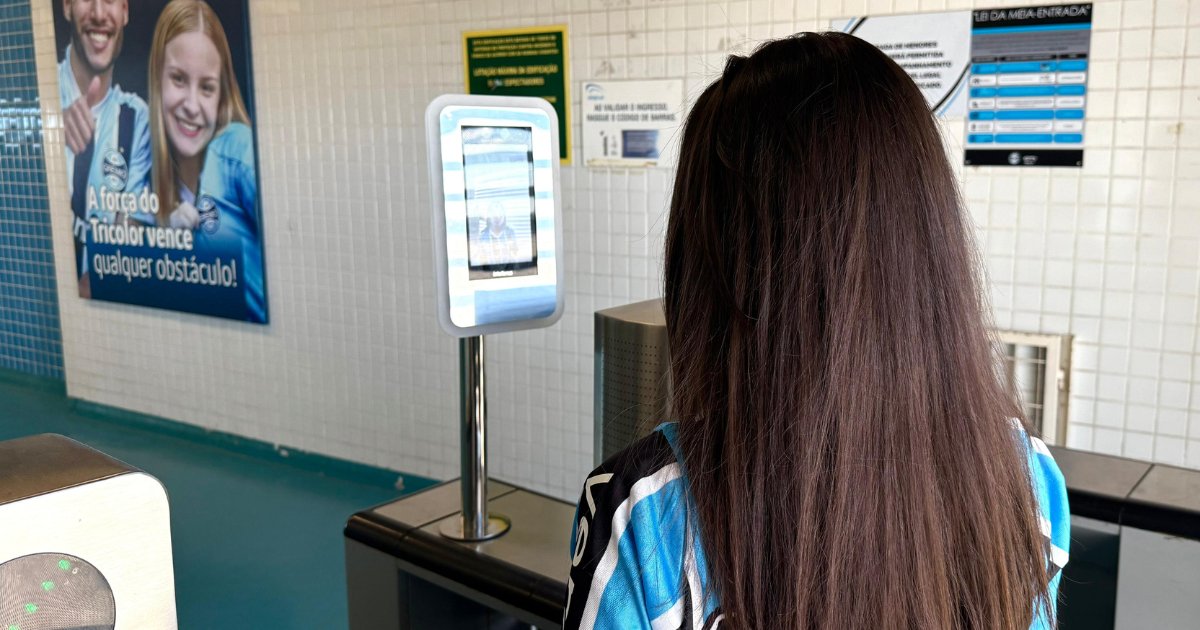 Imagem de uma mulher de costas utilizando um terminal de reconhecimento facial na Arena do Grêmio. Ao fundo, um painel com imagens promocionais do clube e mensagens motivacionais, além de sinalizações informativas nas paredes de azulejo branco (arena grêmio, reconhecimento facial, grêmio)