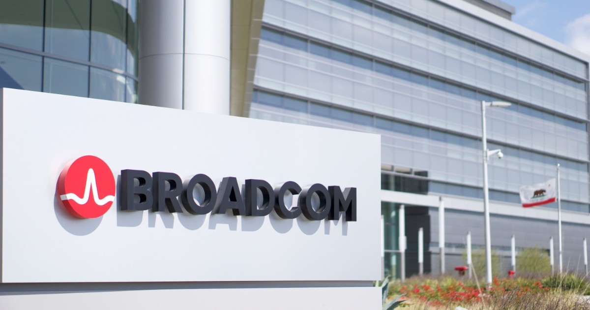 Fachada da sede da Broadcom, com um grande letreiro em primeiro plano exibindo o logotipo da empresa: um símbolo vermelho com uma linha branca ondulada e o nome "Broadcom" em letras pretas. Ao fundo, há um prédio moderno de vidro e metal, com linhas horizontais e janelas espelhadas, além de um céu azul e vegetação na área externa. A composição transmite modernidade e inovação tecnológica (vmware)