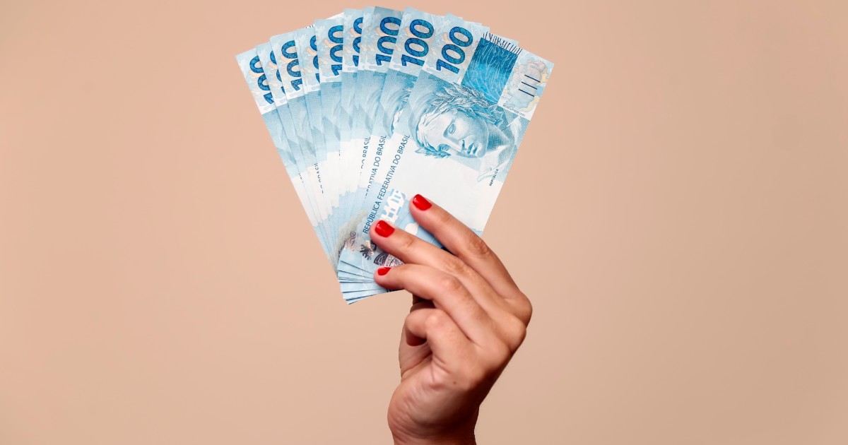 Imagem ilustrativa destacando várias cédulas de 100 reais sendo seguradas por uma mão feminina, representando conceitos como eConsignado, crédito consignado ou finanças pessoais, em um fundo bege uniforme (PaGol)