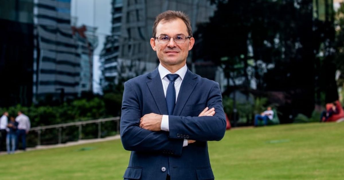 Imagem de Marcos Ferrari, presidente-executivo da Conexis, em um ambiente externo. Ele está usando um terno azul escuro com gravata, de braços cruzados, transmitindo uma postura confiante e profissional. Ao fundo, há um gramado verde, árvores e edifícios modernos, criando um cenário corporativo ao ar livre