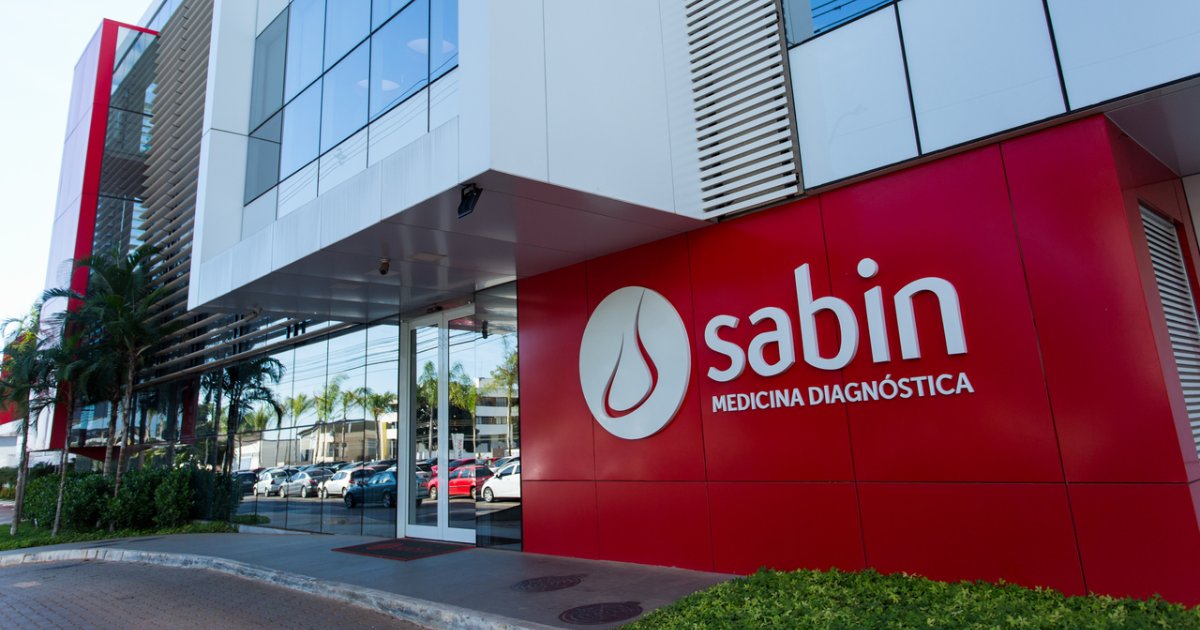 Fachada do laboratório Sabin Medicina Diagnóstica com design moderno, destacando a marca em letras brancas sobre uma parede vermelha, com estacionamento e paisagismo ao fundo (nuvem, sabin, medicina diagnóstica, saúde)