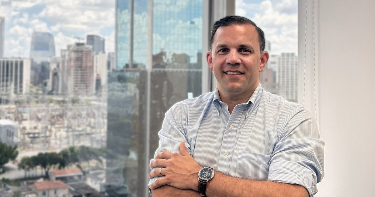 A imagem mostra Agustín Durán, CEO da Seidor Brasil, posando em um escritório com uma grande janela ao fundo, que revela uma vista panorâmica de prédios comerciais em uma cidade moderna. Ele está usando uma camisa social clara e um relógio de pulso, com os braços cruzados e um sorriso confiante. A iluminação natural realça a atmosfera profissional da foto (Agustín Durán, Seidor Brasil, Carlyle)