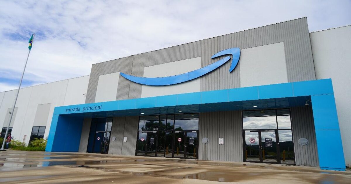 Imagem do novo centro de distribuição da Amazon Brasil localizado em Cajamar. O edifício é grande e moderno, com uma fachada cinza e detalhes em azul. O logotipo da Amazon, com sua seta característica, está em destaque no topo da construção. Na entrada principal, há portas de vidro e uma cobertura azul clara. O piso molhado reflete parte da estrutura, enquanto o céu nublado e a bandeira do Brasil ao fundo complementam a cena