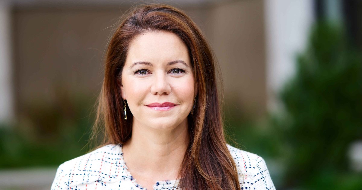 Retrato de Amy Fowler, gerente comercial geral da Pure Storage. Ela está sorrindo levemente, com cabelos castanhos longos e lisos. Veste uma blusa estampada clara e brincos discretos. O fundo desfocado apresenta uma paisagem externa com elementos verdes, destacando seu rosto iluminado (atualiza)