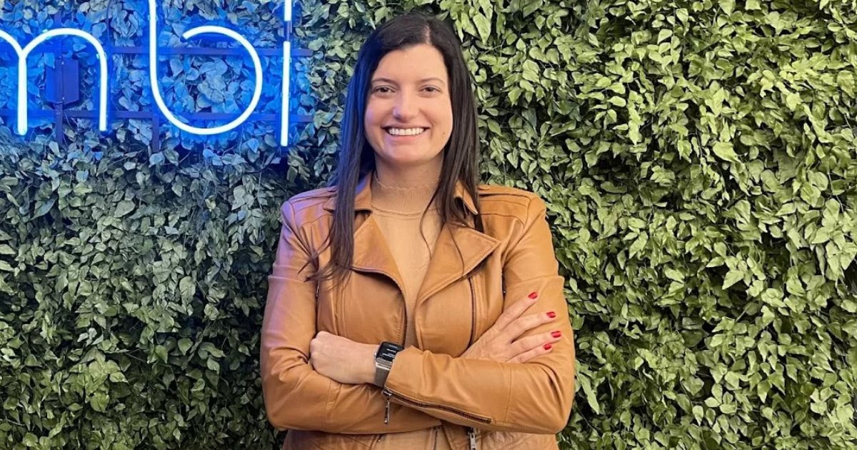 Retrato de Carolina Cabral, CEO da Nimbi, sorrindo e posando com os braços cruzados. Ela veste uma jaqueta de couro marrom sobre uma blusa bege. O fundo é composto por um painel verde com folhagens artificiais e um letreiro neon azul, criando um ambiente moderno e sofisticado (Carolina Cabral, Nimbi OutSystems)