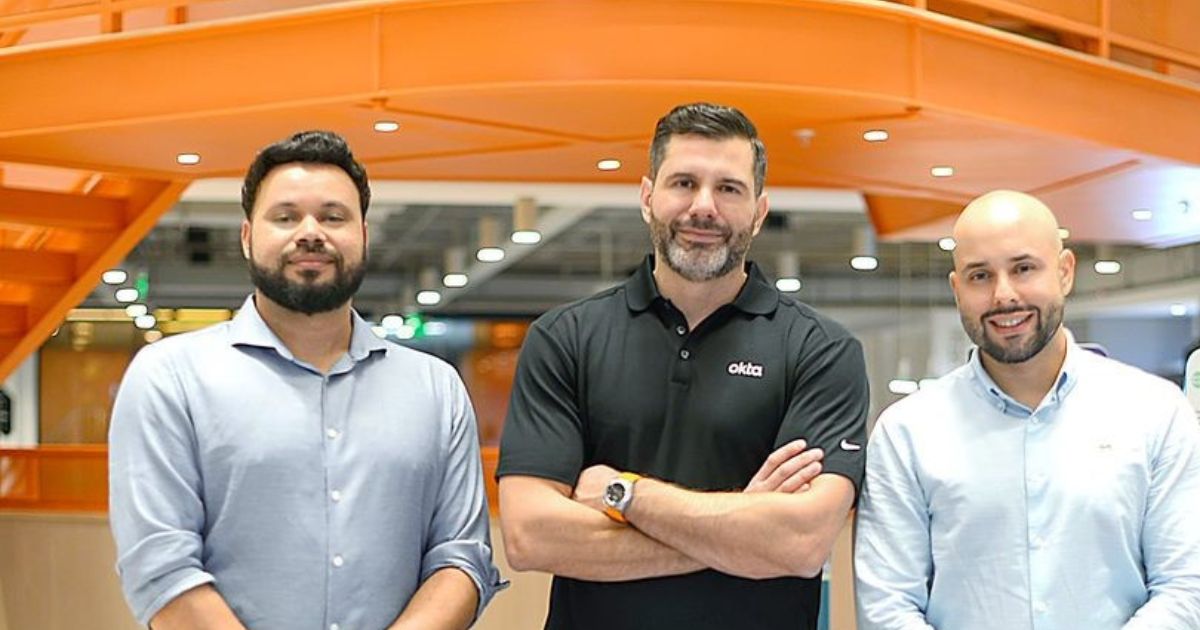 Três homens posam juntos em um ambiente corporativo iluminado com detalhes em laranja no fundo. Da esquerda para a direita: David Junior, cofundador e VP de Operações da Okta, Vini Lima, Account Executive da Okta, e Guilherme Penhalver, cofundador e VP de Vendas e Marketing da Okta.