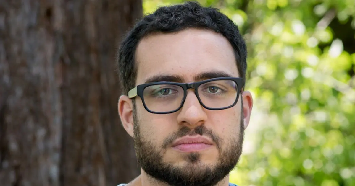 Retrato de um homem com barba, cabelo curto e óculos de armação preta, posando ao ar livre com um fundo desfocado de árvores e folhagem verde. Ele tem uma expressão neutra e está olhando diretamente para a câmera.